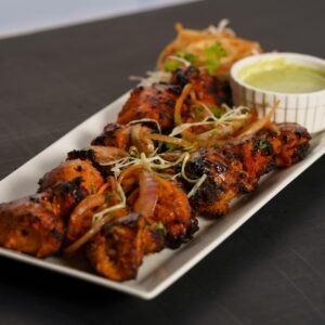 A flavorful chicken tikka dish served with a side of green chutney and garnishes on a white platter.