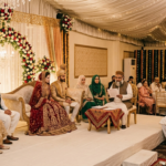 A traditional Islamic Nikah ceremony taking place on an elegant, elevated stage under a beautifully draped white marquee tent with warm fairy lights and chandeliers. In the center, a Maulana with a grey beard, wearing a traditional brown waistcoat and white kurta, sits at a low wooden table reading the marriage contract. To his right, the bride in a rich, heavily embroidered maroon and gold lehenga, and the groom in a gold sherwani with a traditional turban, sit on a plush beige sofa. Family members in formal ethnic attire are seated on both sides of the stage witnessing the ritual.